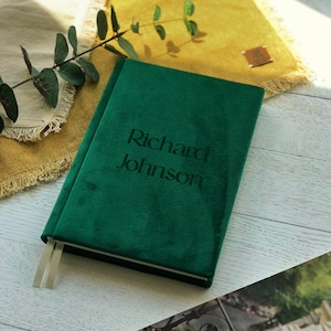 Può includere: Un diario di velluto verde scuro con il nome "Richard Johnson" impresso sulla copertina. Il diario ha un segnalibro a nastro ed è appoggiato su una superficie di legno di colore chiaro. Un rametto di verde e un panno giallo sono sullo sfondo.