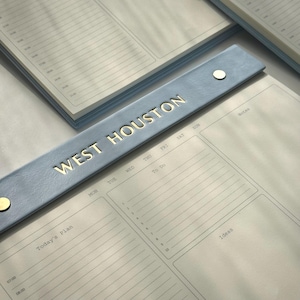Może przedstawiać: Jasnoniebieski planer ze złotym napisem "WEST HOUSTON" na okładce. Planer zawiera sekcje "Today's Plan", "To Do" i "Ideas". Strony są w linie i mają przedziały czasowe od 07:00 do 21:00.