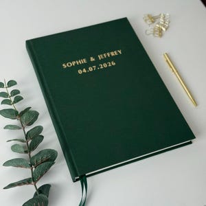 Może przedstawiać: Ciemnozielony notatnik w twardej oprawie ze złotym napisem "SOPHIE & JEFFREY 04.07.2026". Złoty długopis i spinacze do papieru leżą obok notatnika. Widoczna jest zielona wstążka jako zakładka.