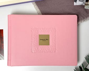 May include: A pink photo album with a textured cover. The album features an embossed floral design around a gold-colored square with the text "George & Alice" and the date. The album is closed and lies flat.