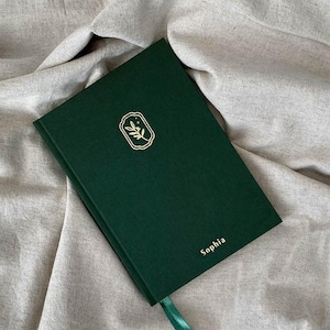 May include: A dark green journal with a gold emblem of a sprig of leaves and the name "Sophia" in gold lettering. The journal has a matching green ribbon bookmark and is set against a neutral fabric background.