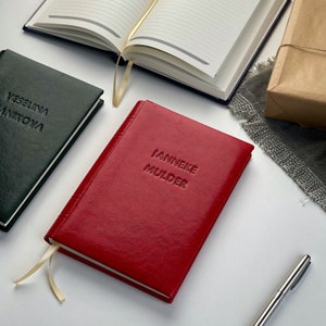 Op de afbeelding: Een rood leren notitieboek met de naam "IANNEKE MULDER" in reli&euml;f op de kaft. Een groen notitieboek met de naam "VESELINA ANIKOVA" is ook zichtbaar. Andere notitieboeken en een pen zijn op de achtergrond.