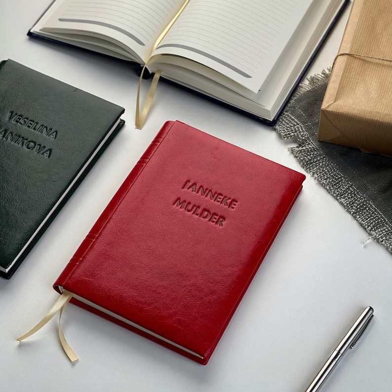 Op de afbeelding: Een rood leren notitieboek met de naam "IANNEKE MULDER" in reli&euml;f op de kaft. Een groen notitieboek met de naam "VESELINA ANIKOVA" is ook zichtbaar. Andere notitieboeken en een pen zijn op de achtergrond.