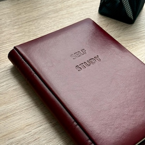 Op de afbeelding: Een bordeauxrood leren notitieboek met de woorden "SELF STUDY" in reli&euml;f op de kaft. Het notitieboek is gesloten en ligt op een lichtgekleurd houten oppervlak. De kaft heeft een getextureerd uiterlijk.