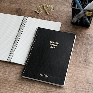 Pode incluir: Um caderno preto com espiral com as palavras douradas "BETTER EVERY DAY". O caderno está sobre uma superfície de madeira ao lado de um caderno aberto com papel quadriculado. Também são visíveis clipes dourados e um porta-canetas.