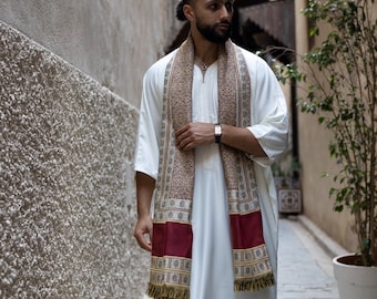 May include: A man wearing a white robe with a long, patterned scarf. The scarf has red, brown, and gold accents. The man is standing in a narrow alleyway.