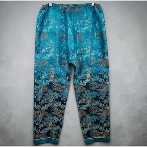 May include: Teal-colored pajama pants with an all-over Asian-inspired landscape print featuring trees, pagodas, and foliage. The pants have a relaxed fit and a white trim along the bottom hem. The fabric appears to be silky.
