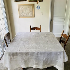 Pode incluir: Uma toalha de mesa retangular branca com padrões florais bordados cobre uma mesa de madeira. Duas cadeiras de madeira estão em ambos os lados da mesa. A sala tem uma paleta de cores neutras com uma janela e uma porta.