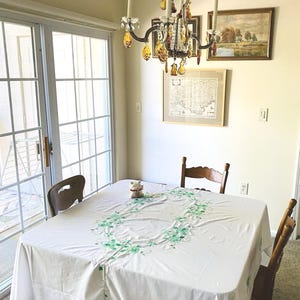 May include: A dining room scene featuring a white tablecloth with a green floral embroidery design. Two wooden chairs are positioned around the table. A chandelier hangs above, and artwork adorns the wall. A small ceramic cat figurine sits on the table.