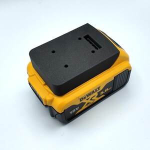 Pode incluir: Um adaptador preto está em cima de uma bateria DeWalt amarela e preta de 18V 5.0Ah. O adaptador tem dois pequenos orifícios e uma ranhura. A bateria tem o logótipo e o texto DeWalt.