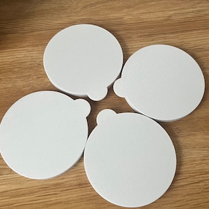 May include: Four white, round coasters with a textured surface and a small tab on one side. The coasters are arranged on a light brown wooden surface. These coasters are designed to protect surfaces from drink rings.