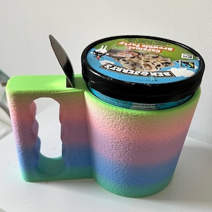 Puede incluir: Un soporte para helado impreso en 3D en un degradado de azul, rosa y verde, con un asa y una ranura para una cuchara. Contiene un bote de helado Ben & Jerry's Caramel Brownie Party, con una cuchara.