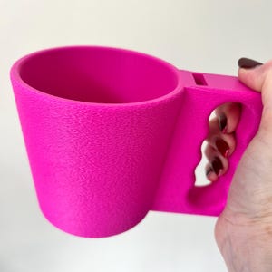 Puede incluir: Una taza rosa brillante con una superficie texturizada. La taza tiene un asa única con un diseño recortado. La taza está sostenida por una mano con esmalte de uñas oscuro. La taza está hecha de un material sólido.