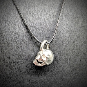 Op de afbeelding: Een zilveren schedel hanger ketting. De schedel hanger is gedetailleerd met oogkassen, een neusholte en een kaaklijn. De hanger is bevestigd aan een zwarte koordketting. De achtergrond is een getextureerd zwart oppervlak.