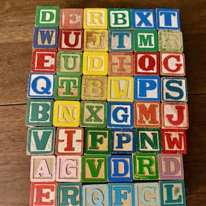 May include: A collection of forty-two square wooden alphabet blocks arranged in a grid. Each block features a single letter printed in various colors including red, blue, green, yellow, and light pink, with some letters showing signs of wear.