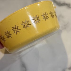 May include: A round yellow bowl featuring a repeating dark brown starburst or snowflake pattern around the exterior side. The bowl has a smooth, glossy finish and a white interior base, showing a clear contrast between the yellow exterior and white.