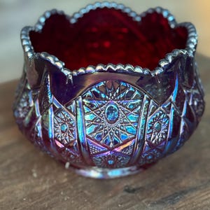 May include: A decorative, iridescent glass bowl with a scalloped rim. The bowl features a deep red interior and an exterior with intricate geometric patterns in shades of blue, purple, and green. The bowl is likely vintage or antique.