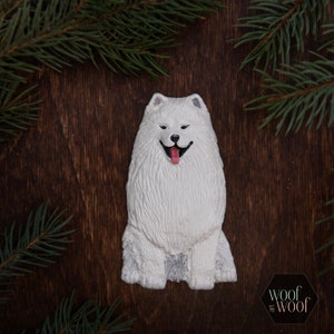 May include: White ceramic ornament of a Samoyed dog with a pink tongue sticking out. The ornament is sitting on a brown wooden surface.