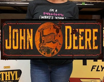 Puede incluir: Un letrero de metal de estilo vintage con fondo negro y texto amarillo que dice "John Deere" con borde rojo. El letrero también presenta una imagen circular de un ciervo rojo con el texto "Plows" en un círculo amarillo.