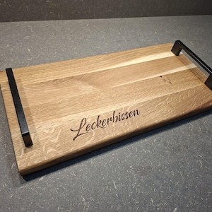 Könnte beinhalten: Ein rechteckiges Serviertablett aus Holz mit einer natürlichen hellbraunen Holzmaserung und zwei schwarzen Metallgriffen an den Seiten. Das Wort Leckerbissen ist in einer kursiven Schriftart auf die Oberfläche des Holzbretts eingraviert.