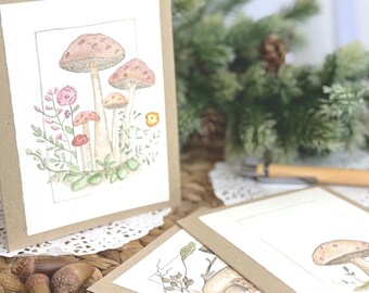 May include: A set of folded greeting cards with a kraft paper border and a white center panel featuring a watercolor illustration of various mushrooms, small pink flowers, and green foliage enclosed within a thin rectangular frame.