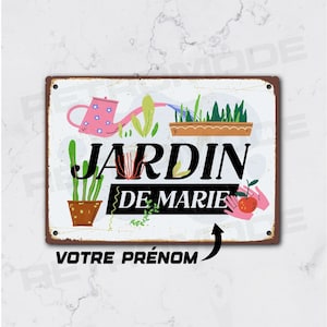 May include: A white metal sign with a rusty brown border. The sign features a colorful illustration of a watering can, potted plants, and a flower box. The text on the sign reads "JARDIN DE MARIE" with a space for a name below.
