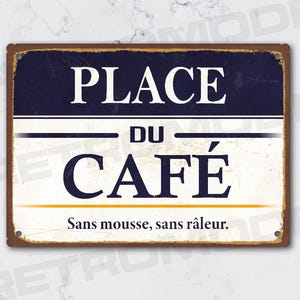 Peut inclure: Panneau métallique rectangulaire à finition vieillie. Il affiche en blanc les mots « PLACE DU CAFÉ » sur fond bleu marine et crème. Le texte « Sans mousse, sans râleur » est en dessous.