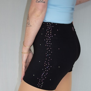Op de afbeelding: Zwarte sportbroek met een sprankelend, gestippeld ontwerp aan de zijkant. De shorts zijn hoog getailleerd en nauwsluitend. Een lichtblauwe top is zichtbaar op de achtergrond. Het woord "Esther" is op de arm getatoeëerd.