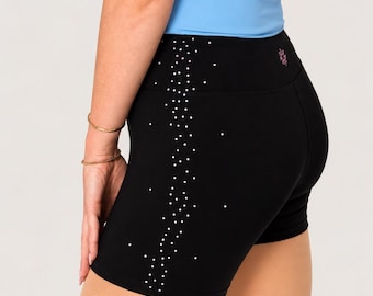 May include: Black athletic shorts featuring a vertical side stripe of scattered white rhinestones and a small pink logo on the back waistband, paired with a light blue tank top and sheer tights embellished with individual rhinestones.
