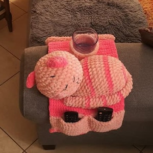 Könnte beinhalten: Ein gemütliches Wohnzimmer mit einem grauen Ecksofa und einem rosa-weiß gehäkelten Organizer. Der Organizer hält ein Glas lila Flüssigkeit und zwei Fernbedienungen. Ein runder, rosa gehäkelter Kissen ist befestigt. Der Raum hat warmes Licht.