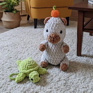May include: A living room with a mustard armchair, a white rug, and a wooden bookshelf. A crocheted bear with a pumpkin hat and a green turtle are on the rug. Plants and a floor lamp create a warm atmosphere. The room has a cosy feel.