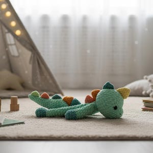 May include: A light green crocheted dinosaur toy with colorful accents of orange, yellow, and blue. The handmade stuffed animal is lying on a light-colored carpet in a child's room. A small teepee and wooden blocks are in the background.
