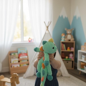 May include: A light green crocheted dinosaur plush toy with blue, orange, and yellow accents. The handmade stuffed animal is held in front of a person in a child's bedroom, with a teepee and wooden bookshelf in the background.