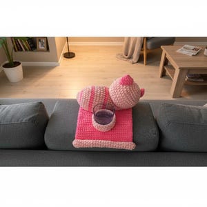 May include: A pink crocheted pillow and teapot set with a matching pink blanket and a glass on a gray sofa. The pillow has pink and white stripes. The teapot has a small handle and a lid.
