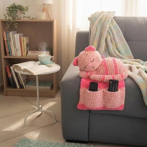 May include: A pink and striped cat-shaped remote control holder rests on a gray sofa. The handmade holder has two pockets for remotes. A cozy blanket and a bookshelf with books are in the background.