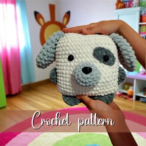 May include: A handmade crochet dog with a square body, gray ears, and a gray nose. The dog has black button eyes and a gray spot on its head. The text "Crochet pattern" is written in cursive at the bottom of the image.