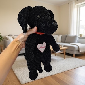 Könnte beinhalten: Ein schwarzer Plüschhund mit rotem Halsband und einem herzförmigen Aufnäher auf der Brust. Auf dem Herzen ist das Wort "Merci" gestickt. Der Hund wird vor einem Wohnzimmer gehalten.