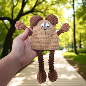 May include: A handmade, plush toy in the shape of a cartoon mouse. The toy is primarily light brown with darker brown accents on the ears, arms, legs, and feet. It has white eyes, a black nose, and a small, stitched mouth. The toy is held in a hand.