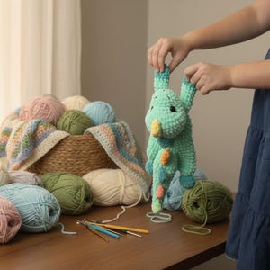 May include: A handmade, light green crochet dragon with yellow and orange accents is held up. A basket filled with yarn in various colors, including pink, blue, and green, sits on a wooden table. Crochet hooks and yarn are also visible.