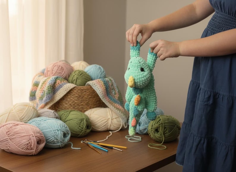 May include: A handmade, light green crochet dragon with yellow and orange accents is held up. A basket filled with yarn in various colors, including pink, blue, and green, sits on a wooden table. Crochet hooks and yarn are also visible.