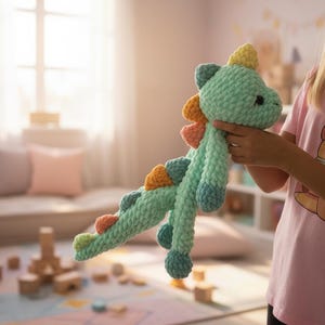 May include: A handmade, light teal crochet dinosaur toy with colorful accents. The dinosaur features a yellow crest, orange and pink spines, and blue feet. The plush toy is held up in front of a window.