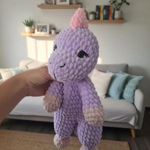 May include: A handmade, plush dinosaur toy in shades of lavender and pink. The soft, textured toy has a pink horn, black eyes, and light pink accents on its feet and hands. The dinosaur is held in front of a blurred background.