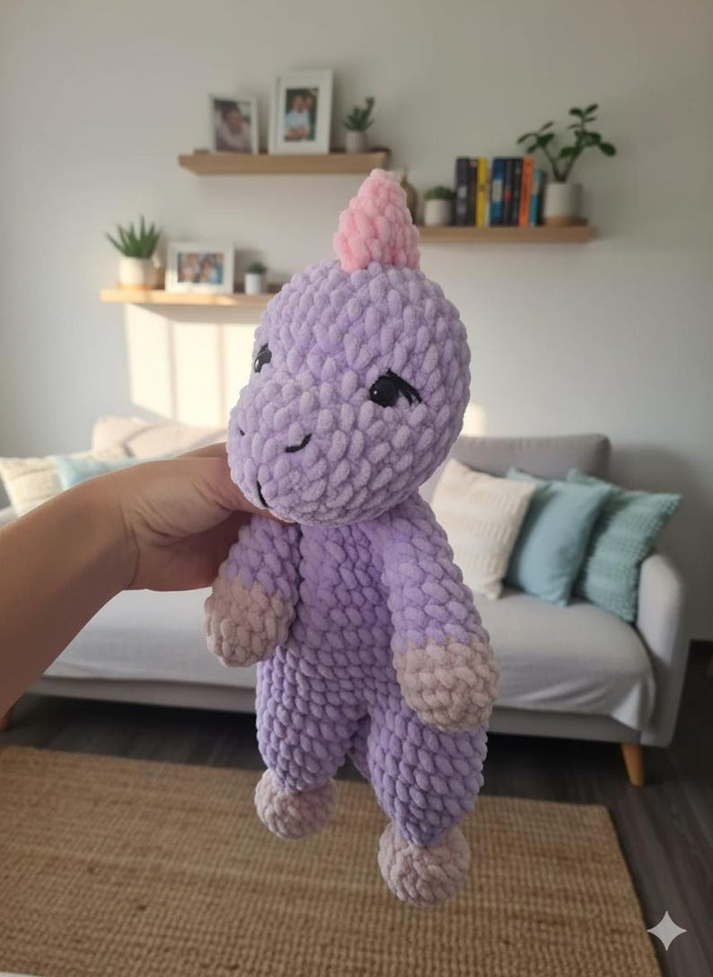 May include: A handmade, plush dinosaur toy in shades of lavender and pink. The soft, textured toy has a pink horn, black eyes, and light pink accents on its feet and hands. The dinosaur is held in front of a blurred background.