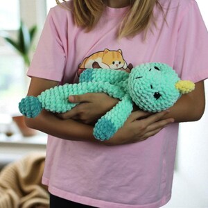 May include: A handmade, teal-colored plush duck toy with a yellow beak and dark eyes. The duck is made of a soft, textured material and is being held by a person wearing a pink t-shirt with a cartoon cat graphic.