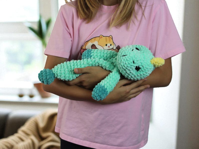May include: A handmade, teal-colored plush duck toy with a yellow beak and dark eyes. The duck is made of a soft, textured material and is being held by a person wearing a pink t-shirt with a cartoon cat graphic.