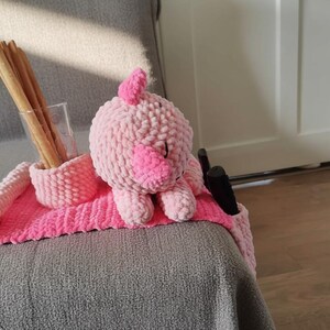 May include: A handmade pink crochet pig-shaped couch arm organizer. The organizer has a pocket for remotes, a holder for a glass of breadsticks, and a pink mat. The pig has a light pink body and a darker pink snout.