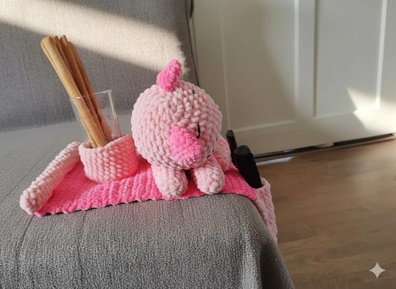 May include: A handmade pink crochet pig-shaped couch arm organizer. The organizer has a pocket for remotes, a holder for a glass of breadsticks, and a pink mat. The pig has a light pink body and a darker pink snout.