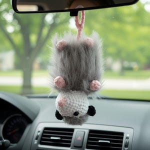 May include: A handmade gray and white crochet car charm shaped like a bat, hanging upside down. The bat has pink accents on its ears and feet, with black eyes and a pink hanging loop. The charm is hanging from a car's rearview mirror.