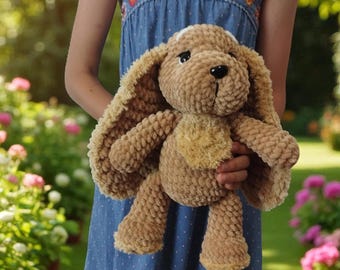 Könnte beinhalten: Ein handgefertigtes Plüschhundespielzeug in Beige- und Brauntönen. Der Hund hat lange, schlaffe Ohren, einen kleinen beigefarbenen Fleck auf dem Kopf und einen helleren beigefarbenen Fleck auf der Brust. Das Spielzeug wird vor einem blauen Kleid mit Blumenmuster gehalten.