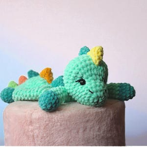 May include: A handmade, plush dinosaur toy in shades of teal, green, yellow, orange, and blue. The dinosaur has a friendly expression with black eyes and is lying on a pink surface. The toy is made of soft, textured yarn.